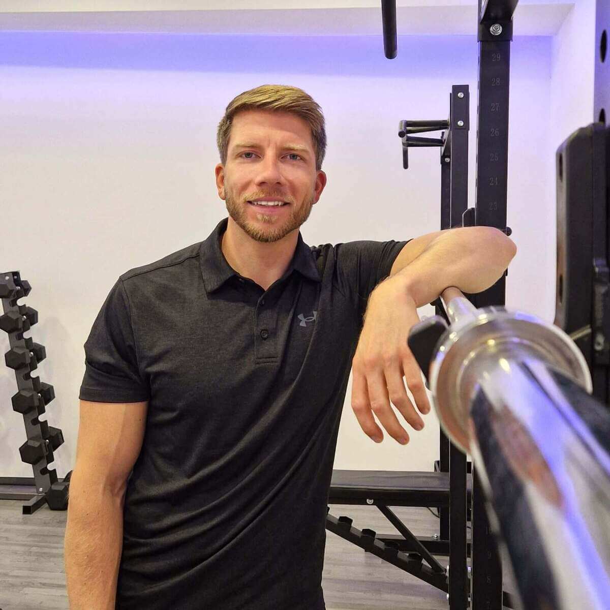 Personal Training in 1010 Wien. Hier sieht man Sport- & Ernährungsiwssenschaftler und Personal Trainer Martin Fiedler im Kraftraum an der Langhantel lehnen, welche auf dem Rack liegt. Er schaut freudig in die Kamera